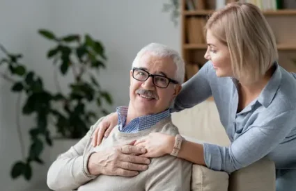Restful senior man on couch holding hand of his young blonde daughter standing near by and embracing her father
